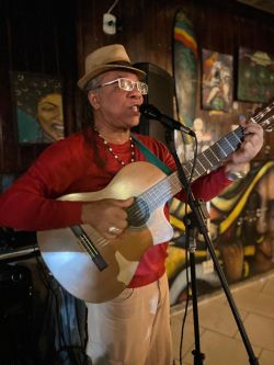A figura mostra Antônio Carlos Mariano, um homem negro, idoso, usando chapéu, óculos de grau, uma guia no pescoço e vestindo uma camisa vermelha, tocando um violão e cantando no microfone.